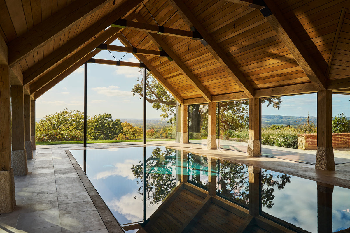 An indoor infinity pool in a modern timber-framed pavilion with a high wooden roof, overlooking a scenic outdoor landscape with trees and hills under a blue sky.